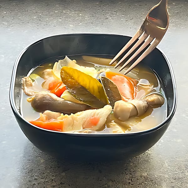 A black bowl of Thai soup with vegetables, mushrooms, and a fork resting inside.